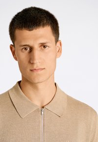 Beige collared tröja med dragkedja, med ett stickat mönster och en slim fit, mot en enfärgad ljus bakgrund.