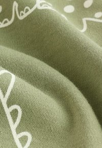 Tissu en coton vert olive, ornée de motifs abstraits blancs. Texture lisse avec un tissage légèrement extensible, montrant des plis et des rides.