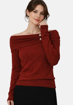 Femme portant un pull rouge rouille à col bateau orné de boutons en perles, posant avec une main près du menton sur un fond uni.