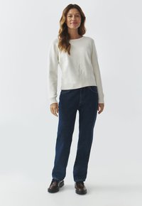 Suéter blanco de punto con puños y dobladillo acanalados, con un sutil diseño en relieve. Jeans rectos de mezclilla azul marino. Zapatos marrones.