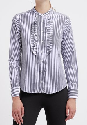 Mujer con camisa de manga larga a rayas verticales en blanco y negro con detalle de volantes en el frente y pantalones negros.