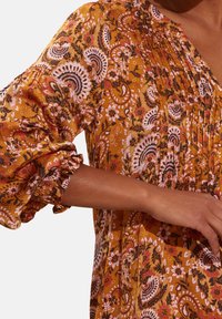 Blusa floreale gialla con design a pieghe, maniche a sbuffo elasticizzate e motivi intricati con accenti rosa e neri. Texture di tessuto morbida.
