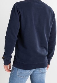 Marineblaue Sweatshirt mit gerippten Ärmeln und Saum, ausgestattet mit einem Rundhalsausschnitt und strukturiertem Stoff. Auffälliges gelbes Logo-Detail an der linken Ärmel.