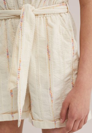 Bež kratke hlače teksture s vertikalnim pastelnim prugama i vezanim strukom, koje nosi osoba s vidljivom desnom rukom koja počiva uz tijelo.