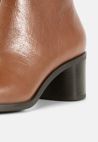 Ruskeat nahkaiset nilkkurit kiiltävällä pinnalla, joissa on musta blokkiolkaiminen korko ja pyöreä varvasmuotoilu. Sileä pinta, jossa vähän yksityiskohtia.