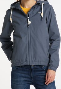 Veste coupe-vent gris foncé, avec une fermeture éclair à l'avant, une capuche ajustable avec cordon et des poignets élastiques. Deux poches latérales sont visibles.