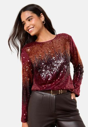 Burgunderfarbiges, mit Pailletten besetztes Langarmoberteil mit einem Farbverlauf von dunkleren zu helleren Nuancen, kombiniert mit eng anliegenden braunen Lederhosen und einem Gürtel.