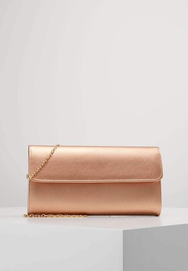 Pochette rectangulaire en or rose avec fermeture à rabat et fine chaîne dorée reposant sur un socle blanc sur un fond gris clair.