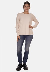 Maglione beige a maniche lunghe con scollatura rotonda, polsini e orlo a costine, indossato sopra jeans blu scuro e stivali neri. Materiale morbido e leggero.