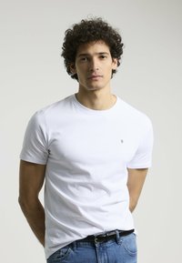Camiseta blanca de manga corta hecha de algodón, con cuello redondo y un pequeño logo en el pecho. Combinada con jeans de mezclilla azul.