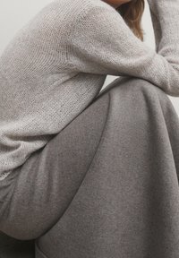 Personne portant un pull en maille lâche gris clair et une longue jupe grise douce, assise avec les bras posés sur les genoux relevés sur un fond neutre.