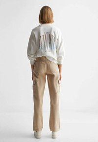 Bílá mikina s pastelovým gradientním nápisem "LATE ALLWAYS CLUB" na zadní straně, doplněná o světle béžové cargo kalhoty a bílé obuv.