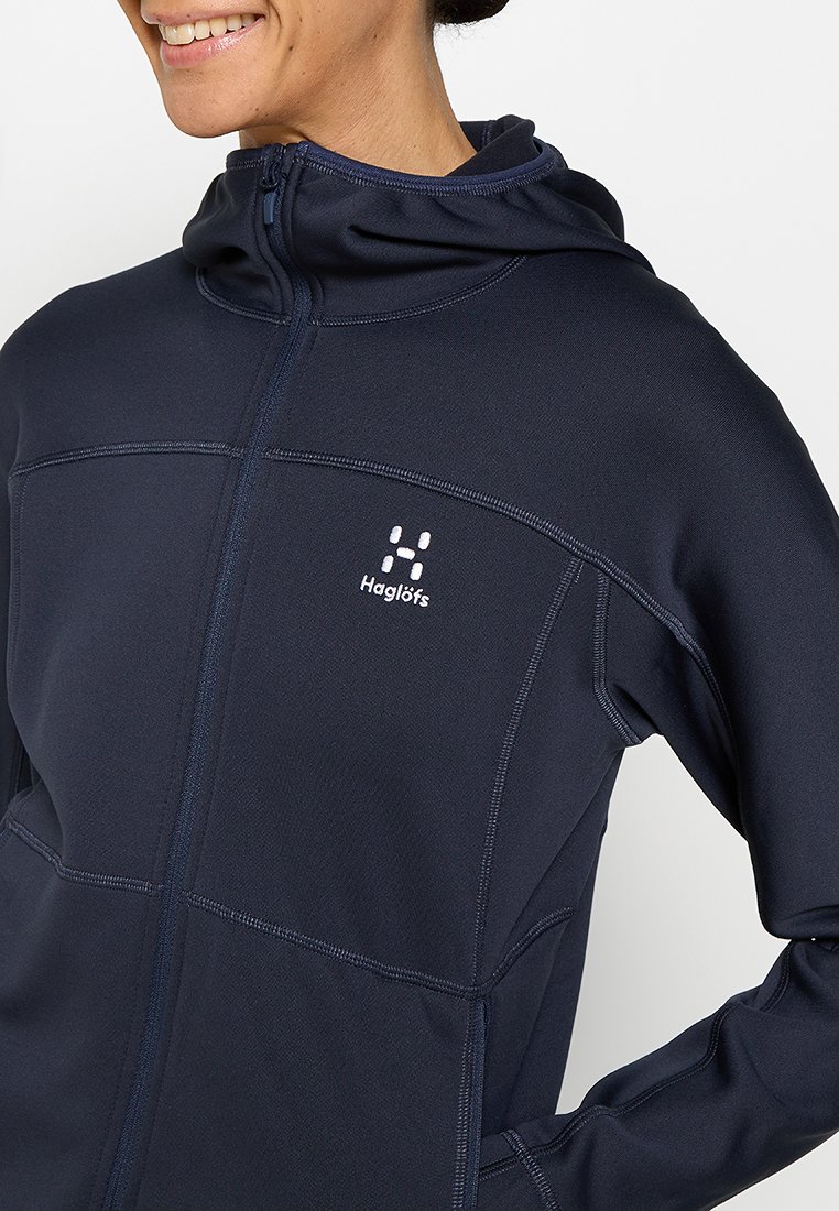 Persona sonriente vestida con una sudadera con cremallera color navy de Haglöfs, con capucha, logo visible en el pecho, mano en el bolsillo, recortada en los hombros y la cintura.