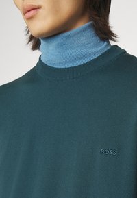 Maglione teal a collo alto realizzato in morbido tessuto con un colletto a costine blu e un discreto logo "BOSS" ricamato sul petto.