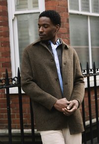 Man wearing brown coat, blue shirt, and beige pants stands with hands clasped in front of a brick building with black iron fence.