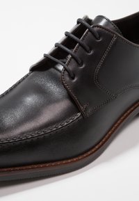 Chaussure en cuir noir à finition lisse, design à lacets, coutures contrastantes et semelle marron. Présente un bout arrondi et des accents minimaux.