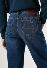Persoon in donkerblauwe denim jeans met achterzakken en een bruine leren merkembleem, gecombineerd met een marineblauwe langemouwtop.