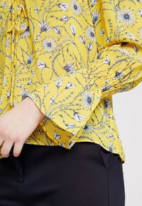 Blouse florale jaune à manches longues, présentant des motifs de fleurs noirs et blancs, des poignets froncés, en tissu léger et avec un ourlet arrondi.