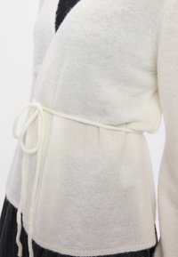 Bílý pletený cardigan s jemnou texturou, s páskem v oblasti pasu a výstřihem do V. Vystaven na tmavém pozadí.