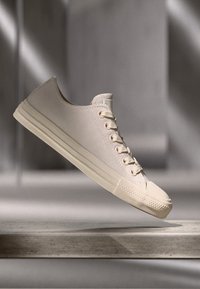 Zapatilla de lona beige con un diseño de caña baja, puntera redonda, seis ojales y suela de goma, mostrada en el aire contra un suave fondo gris.