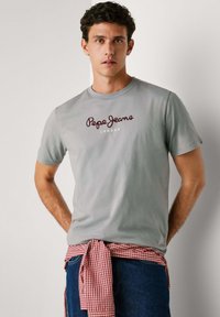 Tricou din bumbac gri cu text "Pepe Jeans London" în nuanță de vișiniu, cu o cămașă casual cu carouri roșii și albe legată în jurul taliei.