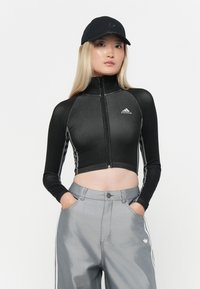 Musta lyhyt vetoketjullinen takki, jossa on korkea kaulus ja valkoiset raidat sivuilla. Yhdistetty vaaleanharmaisiin housuihin, molemmissa on Adidas-logo.