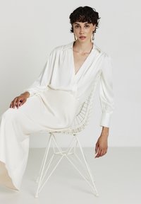 Giovane donna con capelli ricci corti, che indossa una blusa e una gonna di raso bianco, seduta su una moderna sedia di filo bianco su uno sfondo semplice.