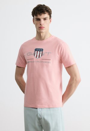 Jeune homme portant un t-shirt GANT rose clair avec un logo en forme de bouclier et le texte « 1949 American Sportswear », associé à un jean bleu clair.
