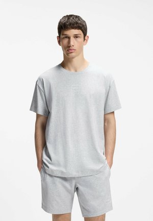 Młody mężczyzna stojący z rękami w kieszeniach, ubrany w jasnoszarą koszulkę Hugo i pasujące szorty na jednolitym białym tle.