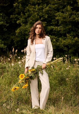 Ung kvinde iført en lysebeige dragt holder visne solsikker og står på en græsmark med tætte grønne træer bag sig.