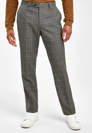 Homme portant un pantalon à carreaux gris, des baskets blanches et une chemise marron à manches longues, debout les mains détendues le long du corps.