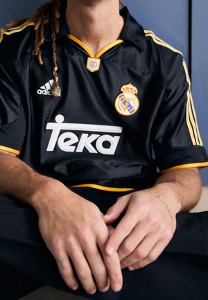 Persoon die een zwart Real Madrid-shirt draagt met gele accenten en Teka-logo, zittend met de handen op zwarte broek rustend.