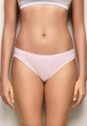 Kobiecy tors w jasnoróżowej bieliźnie z koronkową lamówką i białym, krótko skrojonym topie, stojący na jednolitym tle.