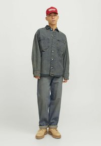 Camicia di denim blu scuro con due tasche sul petto, abbinata a jeans blu chiaro e scarpe beige. Un berretto rosso brillante completa l'outfit.