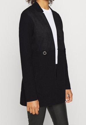Femme portant un blazer noir à manches longues avec des détails de boucles argentées sur un haut blanc et un pantalon noir, debout devant un fond uni.