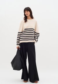 Beige csíkos pulóver fekete vízszintes csíkokkal, fekete, laza nadrággal párosítva. A modell egy nagy fekete táskát tart, amely sima textúrájú.