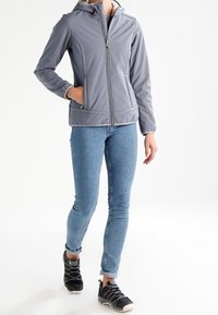 Veste à capuche en softshell grise, avec fermeture éclair et poches latérales, portée sur un jean bleu et des chaussures de randonnée noires.