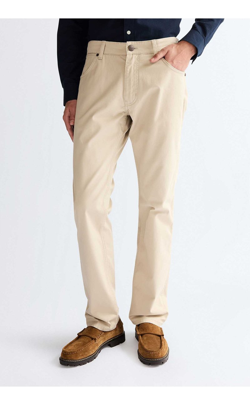 Mann trägt beige gerade geschnittene Hose mit Vordertaschen, kombiniert mit braunen Wildleder-Loafern, steht vor einem einfarbigen Hintergrund.