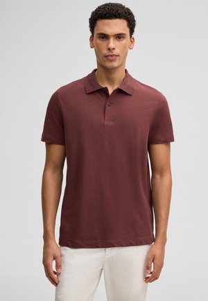 Mann mit lockigem Haar, der ein kurzärmeliges, kastanienbraunes Poloshirt und cremeweiße Hosen trägt, vor einem schlichten grauen Hintergrund stehend.