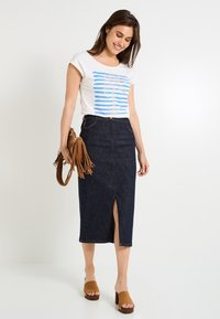 Camiseta blanca con gráfico de palmera a rayas azules, combinada con una falda midi de mezclilla oscura con una abertura en la parte delantera, y sandalias de tacón bloque marrones.