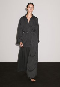 A model walks confidently in a dark striped outfit with a belt, contrasting against a plain light background and dark floor.