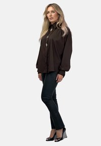 Brązowa koszula zapinana na guziki z wysokim kołnierzykiem i długimi rękawami, zestawiona z dopasowanymi czarnymi spodniami oraz klapkami na koturnie z wzorem w zygzak.