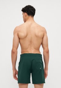 Grønne svømmeshorts har en flat midje, en sidelomme på høyre side, og er laget av et glatt, lett materiale.