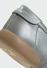 Nærbilde av hælen på en sølvfarget Adidas-sneaker med teksturert, lys brun gummisåle og synlige sømdetaljer.