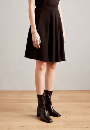 Personne portant une jupe noire mi-longue et des bottines noires à talons, debout sur un sol en bois contre un mur uni.
