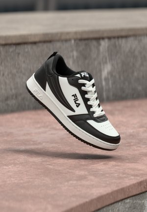 Zapatilla FILA en blanco y negro con cordones blancos flotando sobre una superficie de piedra rosa y un fondo de pared gris.