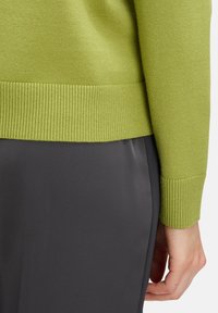 Zöld kötött pulóver bordás aljjal és mandzsettákkal, sima, sötétszürke nadrággal párosítva. A textúrára és a színkontrasztra helyezett hangsúly.