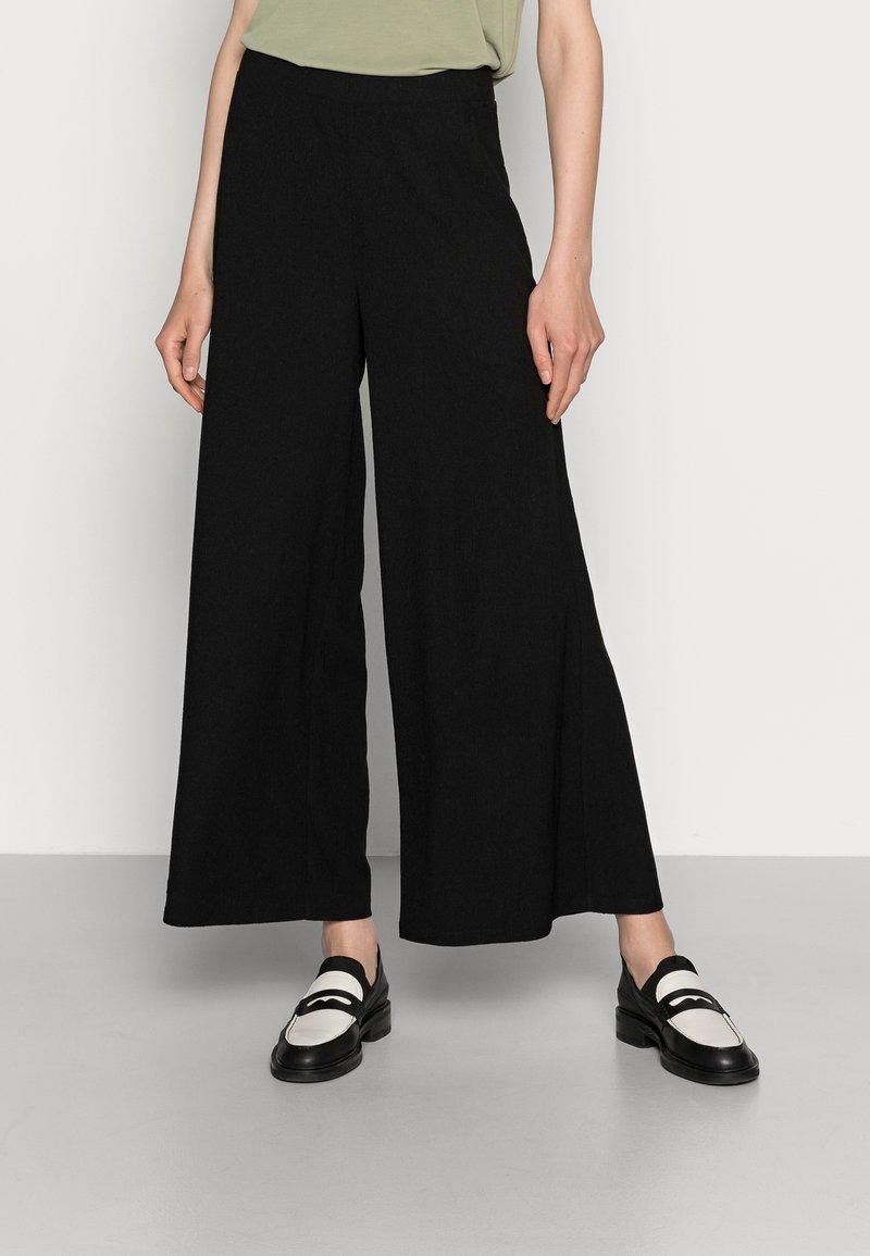 Mannequin portant un pantalon noir taille haute à jambes larges associé à des mocassins noirs et blancs, debout contre un mur uni.
