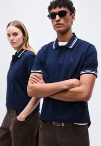 Bărbat cu ochelari de soare și brațele încrucișate și femeie cu mâinile în buzunare, amândoi purtând tricouri polo bleumarin și pantaloni maro, stând pe fundal alb.