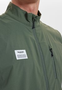 Zelená zipová bunda s vysokým límcem, se bílou nášivkou a dvěma zipovými kapsami. Hladký povrch s lehkým materiálem.